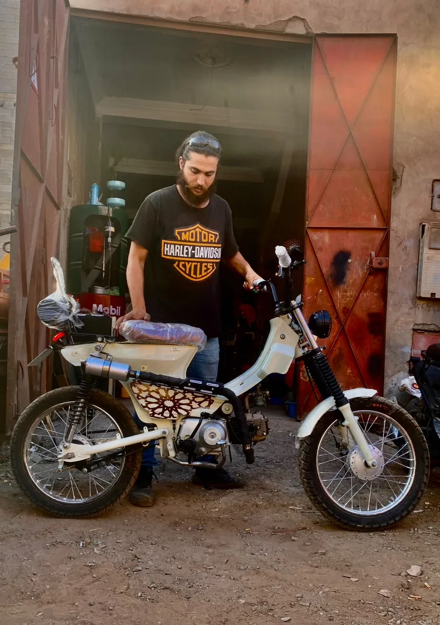 Mouad El Baraka standing with a completed white custom motorcycle at the workshop entrance